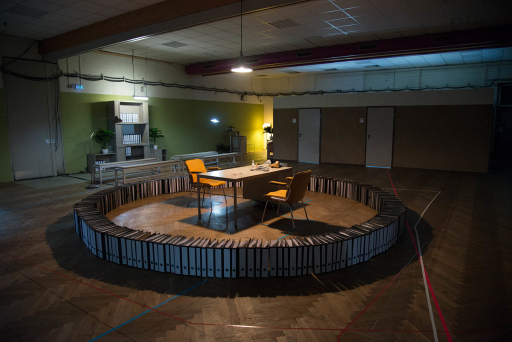 A circle of file folders in a room. Inside the circle is a table with two chairs facing one another. In the background is a large shelf with files.