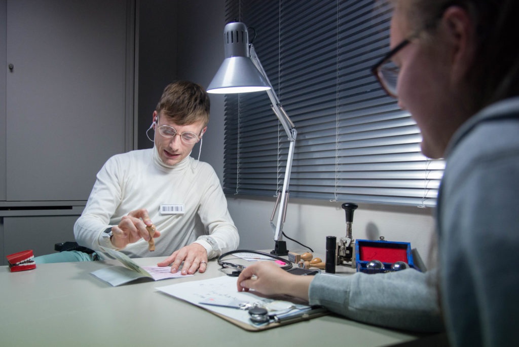 A person with a barcode nametag on their chest stamps a piece of paper at their desk while being lit by a office lamp.