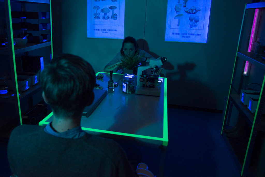 Two people sit across from one another at a table in a dark room. On the table are a microscope, stamps, and other objects.