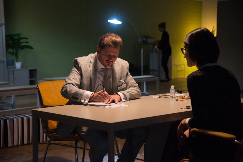 A person in a grey suit sits at a desk and scans a document with a pen while being observed by a person sitting across from them.