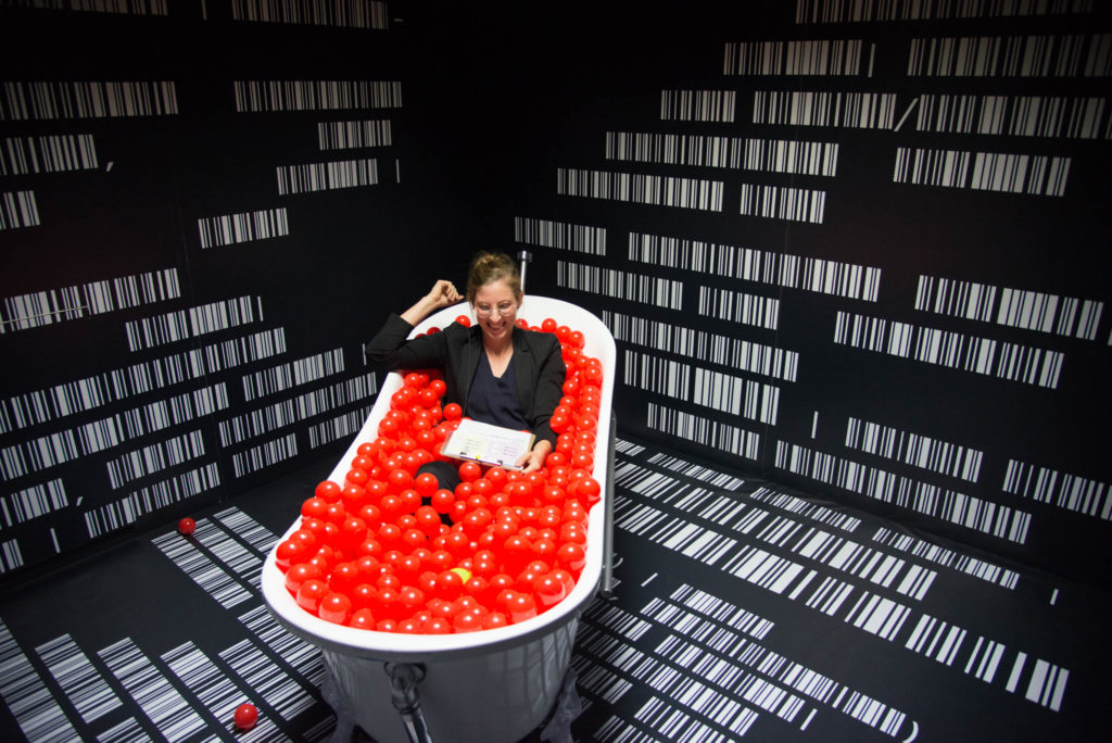 A person with a clipboard is sitting in a bathtub and laughing. The bathtub is full of red plastic balls.