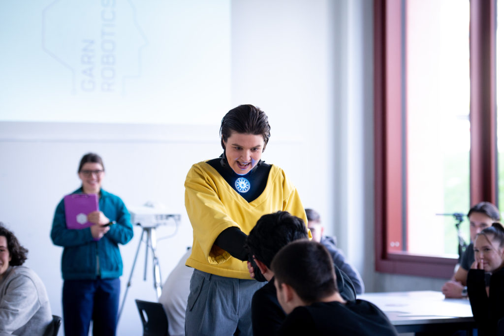 Eine Person mit einem leuchtenden Kreis auf dem Brustbein redet mit einem Schüler in einem Klassezimmer.