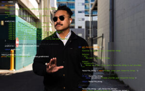 Eine Person mit einer Sonnenbrille steht auf der Straße und hebt die Hand zum Stopzeichen Richtung Kamera. Über dem Foto ist Programmcode eingeblendet.