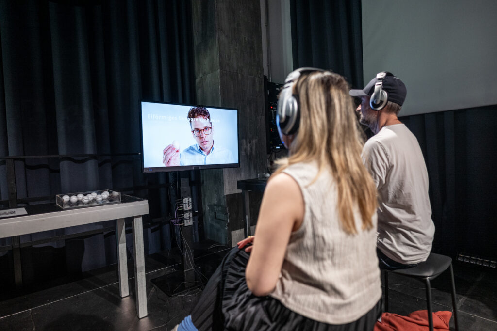 Two people sit in front of a screen with headphones.
