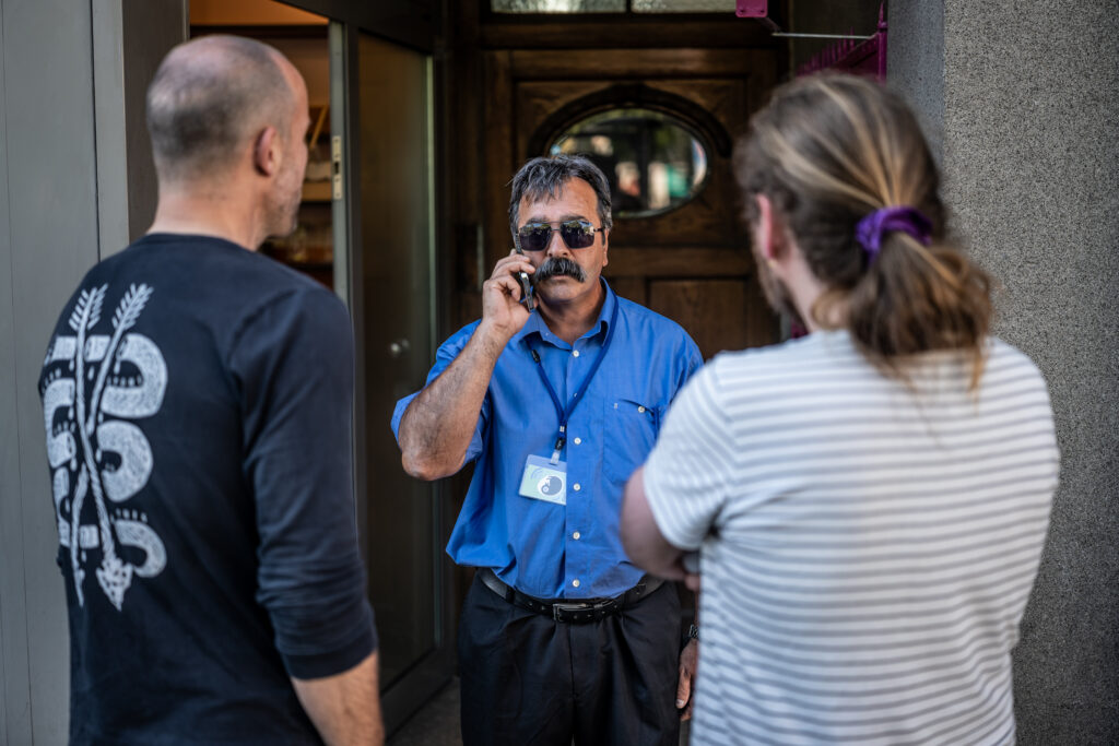 Two people stand in front of a man with a mustache who is talking on the phone.
