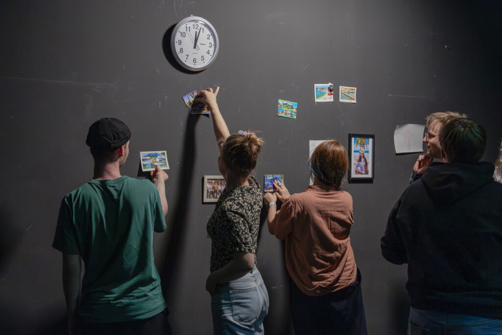 Three people are sticking postcards on a black wall beneath a wall clock.