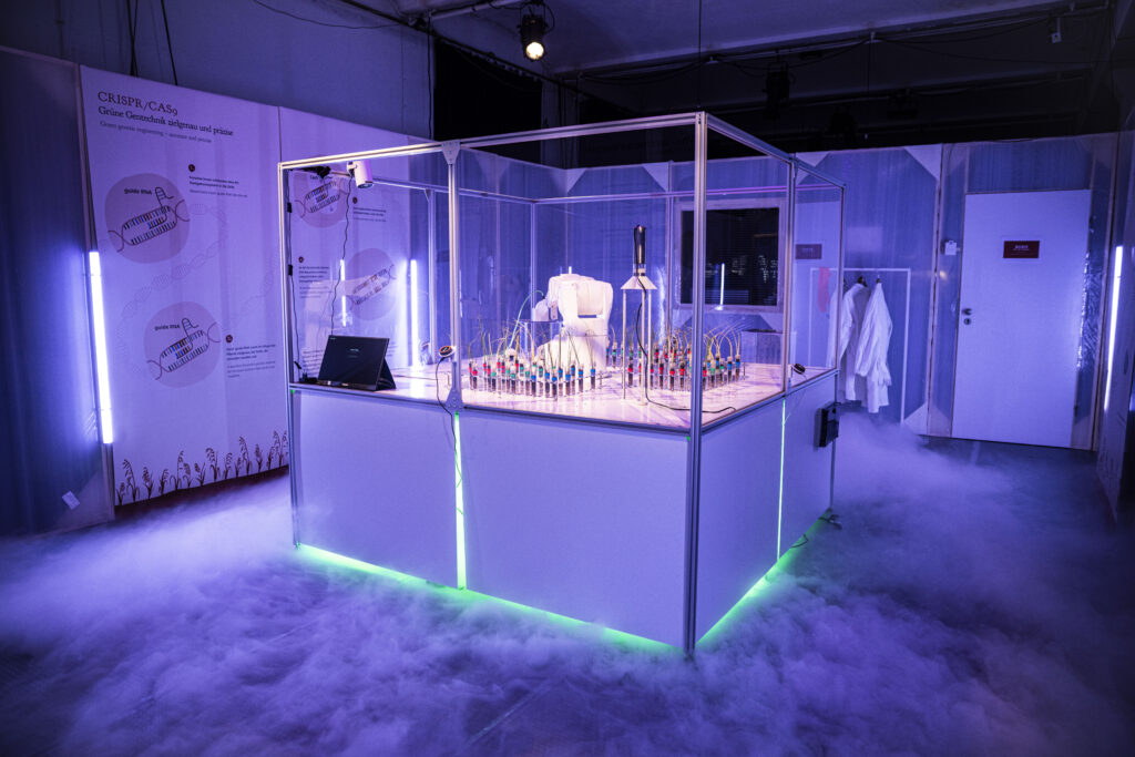 Ein großer Schaukasten mit einem Industrieroboter und Pflanzen in Reagenzgläsern in einem Labor-Ambiente.