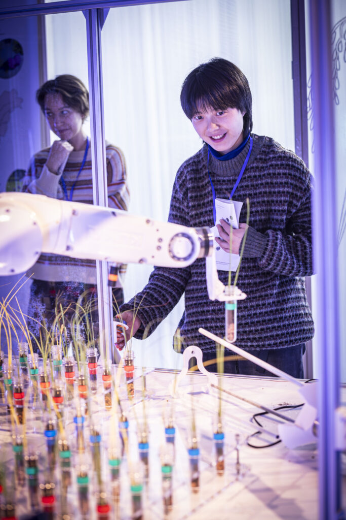 Eine lächelnde Person steht vor einem Tisch mit einem Industrieroboter und Pflanzen im Reagenzglas.