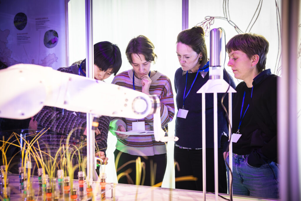 Vier Personen stehen vor einem Industrieroboter und einem Raster mit Pflanzen im Reagenzglas.