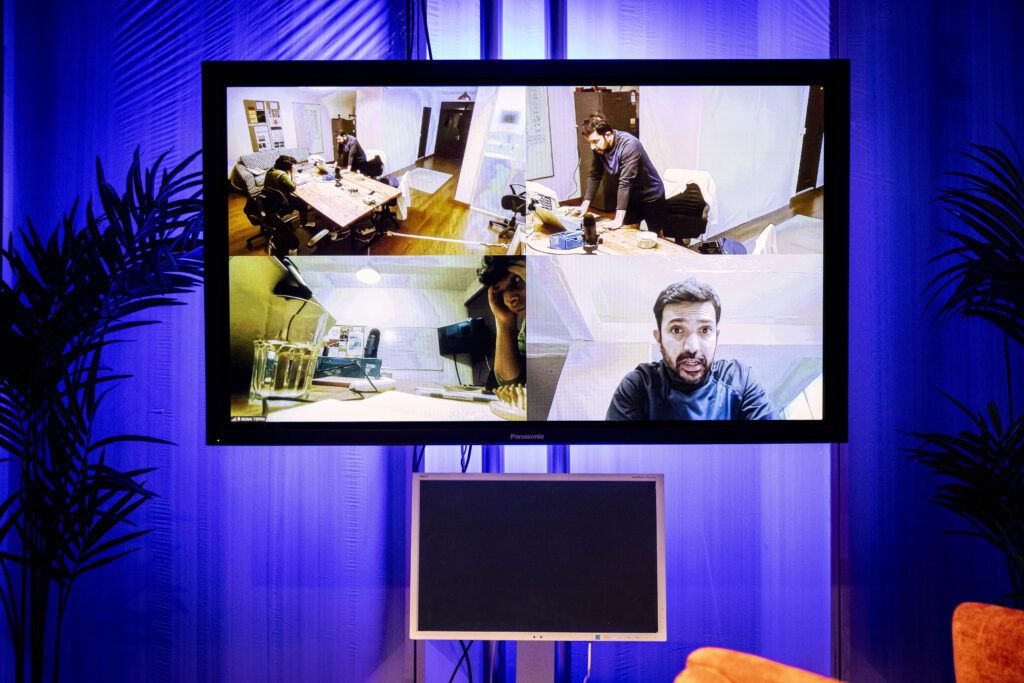 Ein großer Bildschirm mit Split Screen, das zwei Menschen an einem Tisch aus verschiedenen Winkeln zeigt.